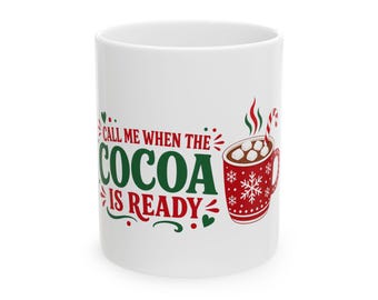May include: A white ceramic mug featuring the red and green text "CALL ME WHEN THE COCOA IS READY" alongside an illustration of a red mug filled with cocoa, marshmallows, and a candy cane, decorated with white snowflakes and green hearts.