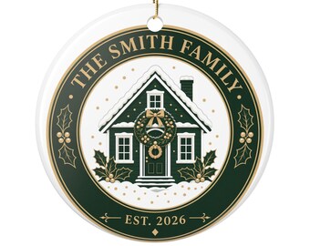 May include: A round white ceramic ornament featuring a dark green circular border with gold accents. The center displays a snow-covered house with a wreath on the door, text reading THE SMITH FAMILY at the top, and EST. 2026 at the bottom.