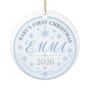 May include: A round white ceramic Christmas ornament featuring a light blue border and snowflake patterns. The text reads Baby's First Christmas, the name Emma in a large script font, and the year 2026. A gold string is attached at the top.