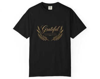 May include: A black short-sleeve t-shirt featuring a gold graphic on the chest. The design includes the word Grateful in script, the text THANKSGIVING 2024, and two symmetrical wheat stalks curving upward to frame the central lettering.