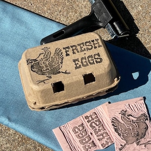 May include: A tan cardboard egg carton featuring a black stamped graphic of a person riding a chicken and the text "FRESH EGGS". A black rubber brayer and two pink linoleum carving blocks with the same design are positioned nearby.