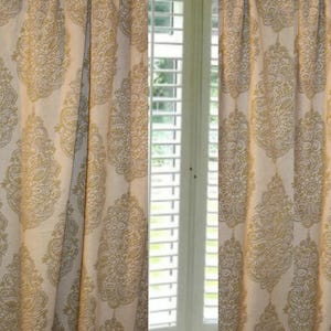 Pode incluir: Um par de cortinas bege com um padrão de damasco dourado. As cortinas estão penduradas em uma janela com persianas brancas.