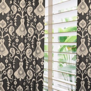 May include: Dark gray and cream-colored patterned curtains with a repeating floral design. The curtains are hanging next to a window with white blinds, and green foliage is visible through the blinds.