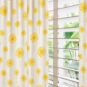 May include: Cream-colored curtains with a repeating pattern of yellow dandelion flowers. The curtains are hanging in front of a window with white blinds, allowing natural light to filter through. The dandelion design adds a touch of nature to the room.