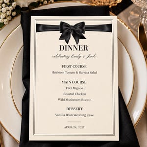 May include: A cream-colored dinner menu card featuring a black ribbon graphic with a bow at the top. The text reads: DINNER, celebrating Emily & Jack, FIRST COURSE Heirloom Tomato & Burrata Salad, MAIN COURSE Filet Mignon, Roasted Chicken, Wild Mushroom Risotto, DESSERT Vanilla Bean Wedding Cake, APRIL 24, 2027.