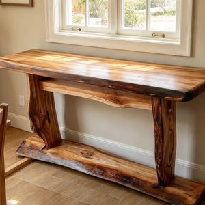 May include: A rectangular console table crafted from natural wood with a live edge finish. The table features a smooth, polished surface with visible grain patterns, supported by two thick, curved wooden legs and a wide base board.