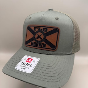 May include: An olive green trucker hat with a tan mesh back and a brown rectangular leather patch on the front. The patch features the text FLO GROWN and a circular seal with crossed rifles. A white sticker on the brim reads 112FPC CHAMP SHAPE OSFM.