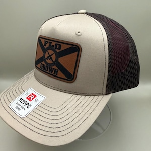 Puede incluir: Una gorra de camionero beige y marrón oscuro con un parche de cuero rectangular en la parte delantera con el texto FLO GROWN y un gráfico circular. Una pegatina blanca en la visera muestra el texto 112FPC, EST 1970, CHAMP SHAPE y OSFM.