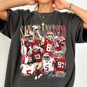 May include: A charcoal gray short-sleeve t-shirt featuring a graphic print of San Francisco football players in red and white uniforms. The design includes the text San Francisco and Football with multiple action shots of athletes.