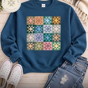 May include: A dark blue crewneck sweatshirt featuring a central graphic of sixteen square quilt blocks arranged in a four-by-four grid. Each block displays a multi-colored star pattern in various shades of pink, green, blue, and yellow.