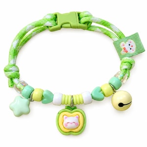Può includere: Un collare in paracord con motivo verde e bianco dotato di fibbia in plastica, una piccola etichetta in tessuto con l'illustrazione di un gatto e varie perline. I ciondoli includono una stella verde chiaro, una mela centrale con muso di gatto e un campanello.