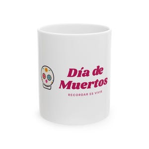 Könnte beinhalten: Ein weißer Keramikbecher mit einer bunten Totenkopf-Illustration auf der linken Seite und dem magentafarbenen Text "Día de Muertos", gefolgt von dem kleineren Text "RECORDAR ES VIVIR", der auf der Seite des zylindrischen Gefäßes aufgedruckt ist.