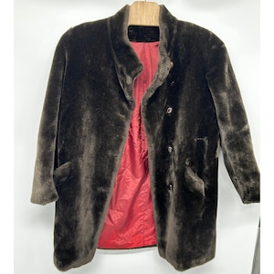 Puede incluir: Un abrigo de piel sintética de color marrón oscuro con cuello alto, mangas largas y un forro interior de color rojo vibrante. La parte delantera del abrigo se cierra con tres botones oscuros e incluye dos bolsillos laterales en una percha.