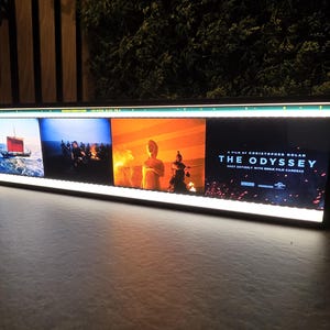 Op de afbeelding: Een rechthoekig, verlicht displaypaneel met vier verschillende filmscènes. Het rechterpaneel toont de tekst: A FILM BY CHRISTOPHER NOLAN THE ODYSSEY SHOT ENTIRELY WITH IMAX FILM CAMERAS, samen met diverse logo's.