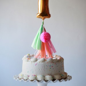 May include: A white cake with colorful sprinkles is decorated with a gold number one balloon, pink and green tissue paper tassels, and a pink honeycomb ball.