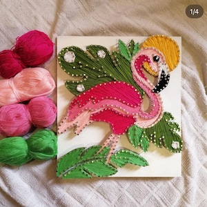 May include: A string art piece on a white board featuring a pink flamingo, green tropical leaves, and a yellow sun, created with colored thread wrapped around metal nails. Four skeins of yarn in pink, light pink, magenta, and green are included.
