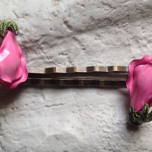 Può includere: Fermaglio decorativo per capelli con due ornamenti a forma di rosa rosa con dettagli verdi. Il fermaglio ha una base in metallo color bronzo. Il fermaglio è posto su uno sfondo strutturato bianco sporco.