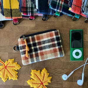 May include: A rectangular plaid zipper pouch featuring a tan, orange, and black pattern with a yellow cord pull. The pouch is displayed alongside a green digital music player with white wired earbuds and two yellow felt maple leaf decorations.