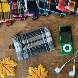 May include: A rectangular zippered pouch featuring a blue, green, and white plaid pattern with a dark green cord pull. The pouch includes a metal D-ring on the side. Nearby are two yellow felt maple leaf cutouts and a green digital music player.