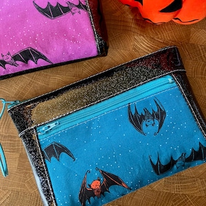 May include: Two rectangular zippered pouches with a Halloween theme. One is purple, the other is blue, both with a bat pattern. The pouches have black trim and zippers. An orange pumpkin is in the background.