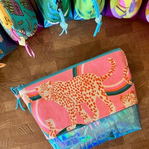 May include: Pink cosmetic bag with a leopard print design and a blue iridescent bottom. The bag features a rainbow and a zipper closure. Other bags with various patterns are visible in the background.