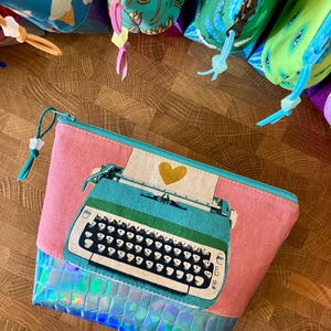 May include: A pink zippered pouch with a teal typewriter graphic and a holographic blue bottom. Other pouches with ice cream and french fry patterns are visible in the background.