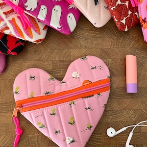 May include: A collection of heart-shaped and rectangular quilted pouches in various patterns, including ghosts, bats, and roses. A pink heart-shaped pouch with an orange zipper and a landscape design is in the foreground, along with a lipstick and white earbuds.