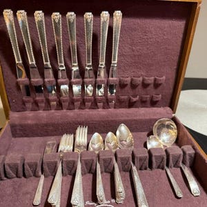 May include: A silver-toned flatware set arranged in a wooden storage case with a dark purple velvet lining. The set includes eight knives with decorative handles, several forks, spoons, and a ladle, all featuring a matching embossed pattern.