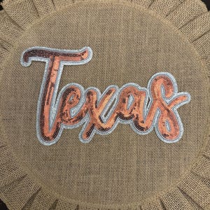 May include: A circular burlap mat with a ruffled edge featuring the word Texas in script lettering. The letters are filled with copper-colored sequins and outlined with a silver border, centered on the textured tan fabric surface.