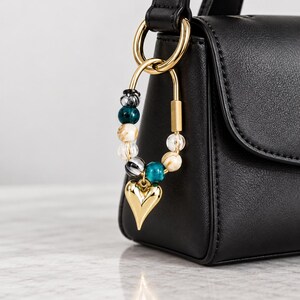 Può includere: Una borsa in pelle nera presenta un ciondolo a forma di U in metallo dorato. Il ciondolo è decorato con una serie di perline nei toni del verde ottanio, bianco e nero, ed è appeso a un anello dorato fissato al manico della borsa.