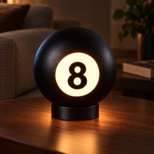 Puede incluir: Una lámpara esférica negra con superficie texturizada que presenta un panel central circular blanco con el número ocho en negro. La lámpara está montada sobre una base cilíndrica negra y emite un brillo cálido desde el centro iluminado.