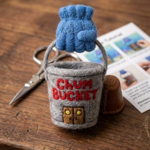 May include: A needle-felted grey bucket with a blue handle, featuring the red text "CHUM BUCKET" and a small brown door with yellow accents on the front. The bucket is crafted from wool and has a textured, soft appearance.