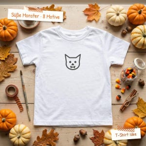 May include: A white short-sleeved t-shirt featuring a central black line-art graphic of a cat face with fangs. The garment is displayed on a wooden surface surrounded by pumpkins, autumn leaves, a paintbrush, and sweets.