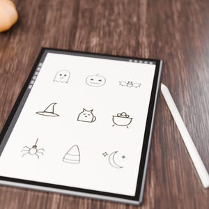May include: A digital tablet with a white screen displaying nine black line-art icons including a ghost, pumpkin, bat, witch hat, cat, cauldron, spider, candy corn, and crescent moon, accompanied by a white stylus pen.