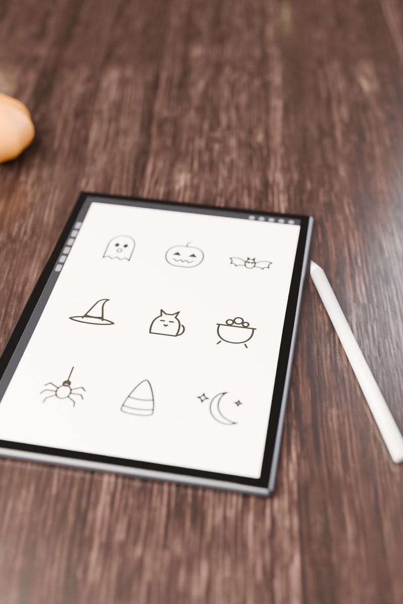 May include: A digital tablet with a white screen displaying nine black line-art icons including a ghost, pumpkin, bat, witch hat, cat, cauldron, spider, candy corn, and crescent moon, accompanied by a white stylus pen.
