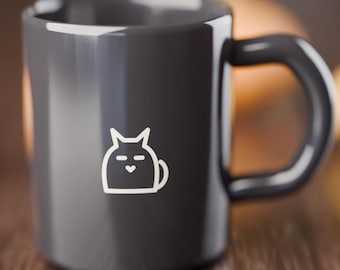 May include: A dark gray ceramic mug featuring a white line art graphic of a stylized cat face with a small heart-shaped nose. The mug has a smooth, glossy finish and a curved handle on the right side, presenting a clean and simple design.