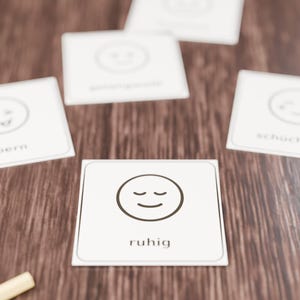 Op de afbeelding: Een set witte vierkante kaarten met zwarte lijntekeningen van gezichtsuitdrukkingen en tekst. De middelste kaart toont een kalm gezicht met gesloten ogen en het woord "ruhig" eronder. Andere kaarten tonen verschillende emoties zoals "schüchtern".