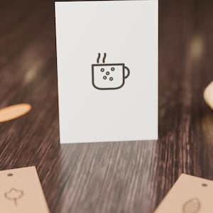 Op de afbeelding: Een witte rechthoekige kaart met een gecentreerde zwarte lijntekening van een dampende mok met cirkelvormige patronen. Twee lichtbruine rechthoekige labels met eenvoudige zwarte lijniconen van een blad en een ovale vorm bevinden zich onder de kaart.