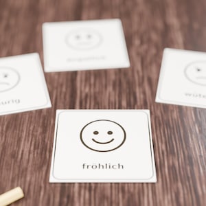 Op de afbeelding: Vier vierkante witte kaarten met afgeronde hoeken, elk voorzien van een zwarte lijntekening van een gezichtsuitdrukking en een Duits woord. De voorste kaart toont een lachend gezicht en het woord fröhlich in zwarte tekst.