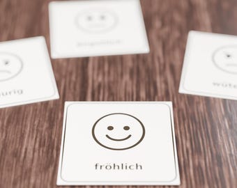 May include: Four square white cards with rounded corners, each featuring a black line drawing of a facial expression and a corresponding German word. The front card displays a smiling face and the word fröhlich in black text.