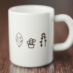 May include: A white ceramic mug featuring three dark brown line art illustrations printed on the front. The designs include a leaf shape with oval cutouts, a small potted cactus, and a tall plant with heart-shaped leaves on a vine.