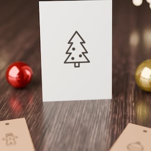 May include: A white rectangular card featuring a centered black line drawing of a Christmas tree with circular ornaments. Two tan rectangular gift tags with simple black line icons of a gingerbread man and a bauble are positioned below.