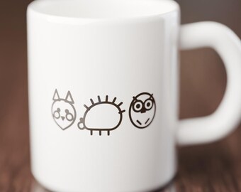 Könnte beinhalten: Ein weißer Keramikbecher mit einer minimalistischen schwarzen Strichzeichnung von drei kleinen Tieren: einer Katze mit spitzen Ohren, einem stacheligen Igel und einer Eule mit rundem Gesicht. Der Becher hat eine glatte, glänzende Oberfläche.