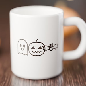 Op de afbeelding: Een witte keramische mok met een zwarte lijntekening van een spook, een pompoen en een vleermuis in een horizontale rij op de voorzijde. De mok heeft een gladde afwerking en een gebogen handvat aan de rechterkant van de beker.