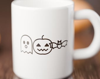 May include: A white ceramic mug featuring a black line art graphic of a ghost, a jack-o'-lantern pumpkin, and a bat arranged in a horizontal row on the front surface. The mug has a smooth finish and a curved handle on the right side.