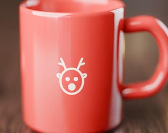 May include: A glossy red ceramic mug featuring a simple white line art graphic of a reindeer face with antlers on the front. The mug has a smooth, rounded handle and a cylindrical body, presenting a clean and minimalist design aesthetic.