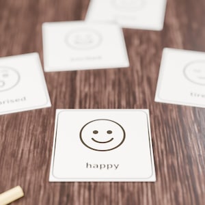 Op de afbeelding: Een set vierkante witte kaarten met zwarte lijntekeningen van gezichtsuitdrukkingen en bijbehorende tekstlabels. De voorste kaart toont een lachend gezicht met het woord happy eronder in een zwart lettertype.