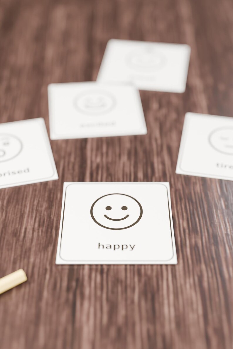 Op de afbeelding: Een set vierkante witte kaarten met zwarte lijntekeningen van gezichtsuitdrukkingen en bijbehorende tekstlabels. De voorste kaart toont een lachend gezicht met het woord happy eronder in een zwart lettertype.