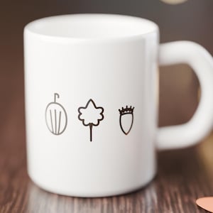 Op de afbeelding: Een witte keramische mok met drie zwarte lijntekeningen op de voorzijde. De iconen tonen een kruisbes, een esdoornblad en een eikel. De mok heeft een glad, cilindervormig lichaam en een gebogen handvat aan de rechterkant.