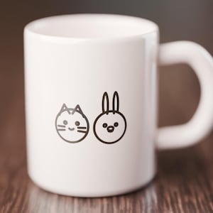 K&ouml;nnte beinhalten: Ein wei&szlig;er Keramikbecher mit einer schwarzen Strichzeichnung eines Katzengesichts und eines Hasengesichts nebeneinander. Der Becher hat einen glatten, zylindrischen K&ouml;rper und einen geschwungenen Henkel auf der rechten Seite.